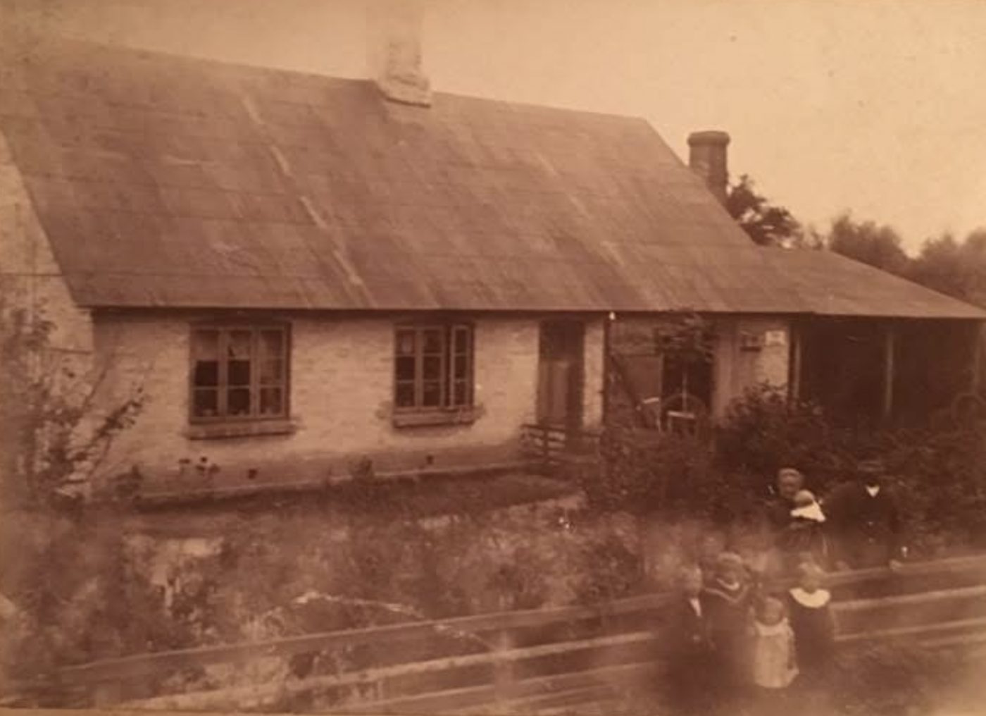 Gammelt foto af Smedehuset: et langt, grundmuret hus med halvtag, en mand og en kvinde med et spædbarn ved buskene og fire børn ved hegnet